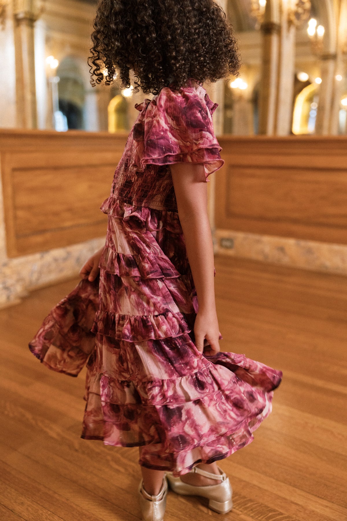 A young girl with curly hair wears the Mini Winnie Dress in Dreamy Roses by Ivy City Co and silver shoes. She twirls on a wooden floor in an elegant room adorned with ornate decor and bathed in golden light.