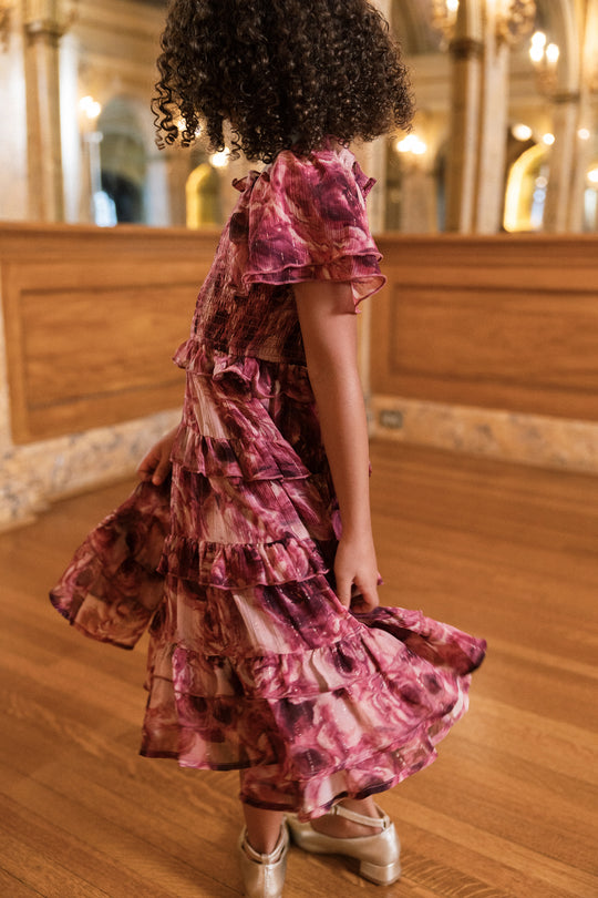 A young girl with curly hair wears the Mini Winnie Dress in Dreamy Roses by Ivy City Co and silver shoes. She twirls on a wooden floor in an elegant room adorned with ornate decor and bathed in golden light.
