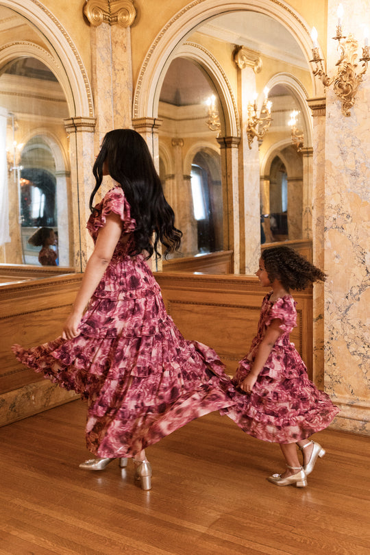 A woman and a young girl in matching floral dresses from Ivy City Co twirl together in an elegant room adorned with marble walls, arched mirrors, and wooden floors. The young girl’s Mini Winnie Dress in Dreamy Roses features charming smocked details, making it a delightful choice for a holiday party dress moment.