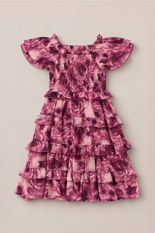The Mini Winnie Dress in Dreamy Roses by Ivy City Co is a pink and purple floral girls’ smocked dress featuring short flutter sleeves, a smocked bodice, and a tiered ruffle skirt, shown laid flat on a beige background. Its charming details make it the perfect holiday party choice and an instant favorite for any festive occasion.