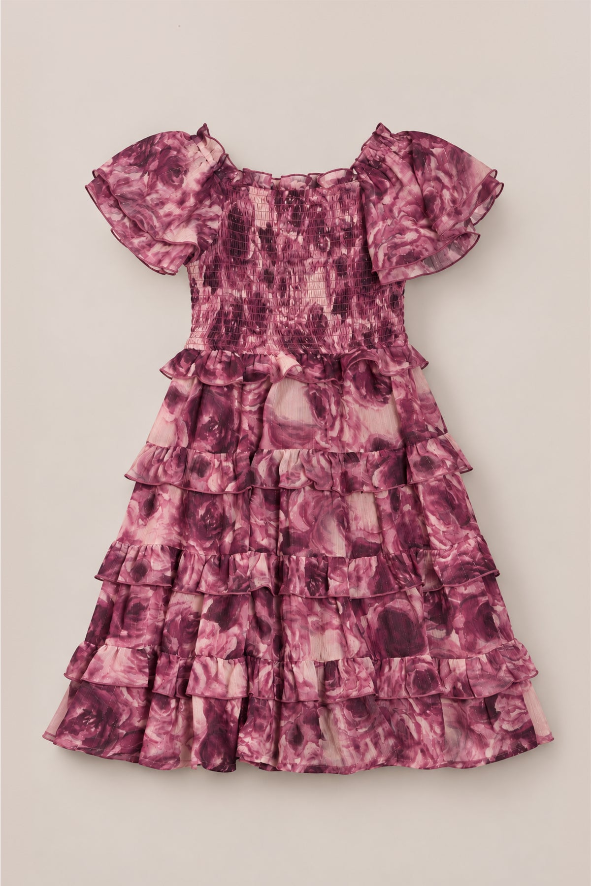 The Ivy City Co Mini Winnie Dress in Dreamy Roses is a charming girls’ dress with short sleeves, a smocked bodice, and a layered ruffle skirt. Decorated in a pink and purple rose floral pattern, this delightful piece is perfect as a holiday party dress and is shown laid flat on a light beige background.