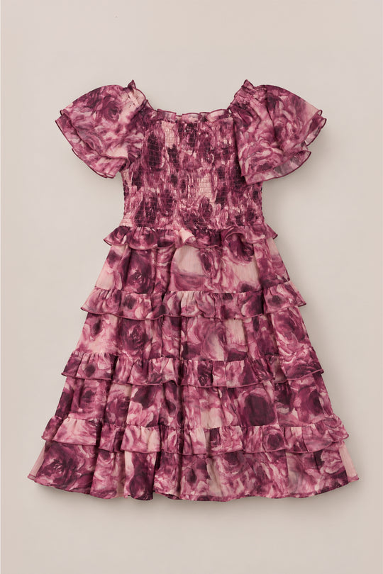 The Ivy City Co Mini Winnie Dress in Dreamy Roses is a charming girls’ dress with short sleeves, a smocked bodice, and a layered ruffle skirt. Decorated in a pink and purple rose floral pattern, this delightful piece is perfect as a holiday party dress and is shown laid flat on a light beige background.