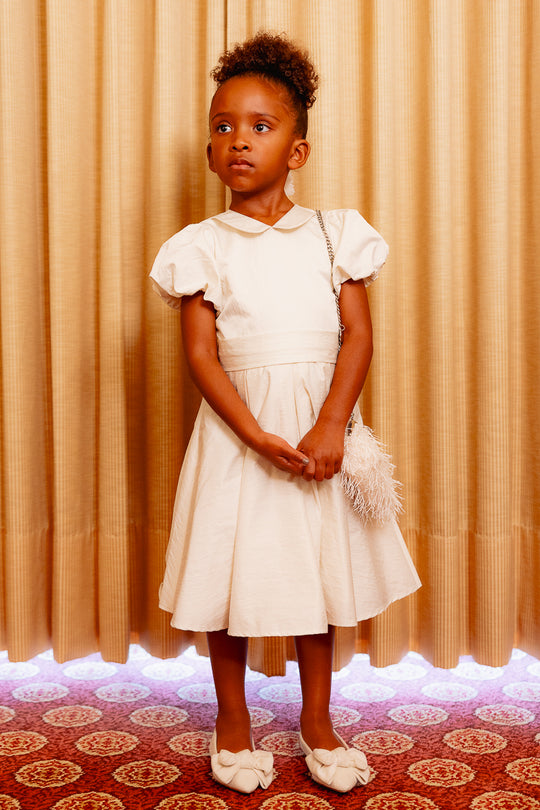 Little girl wearing a white dress standing against a yellow curtain and on a red carpet