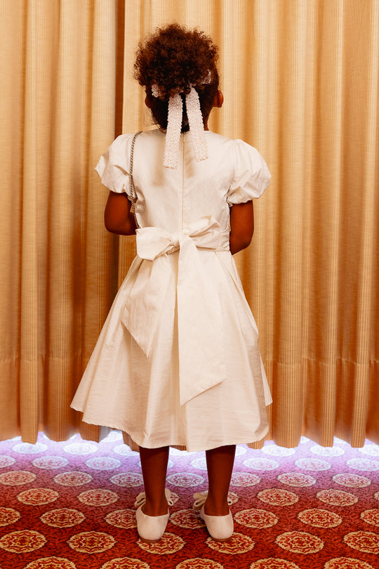 Little girl wearing a white dress standing with her back to the audience against a yellow curtain and on a red carpet
