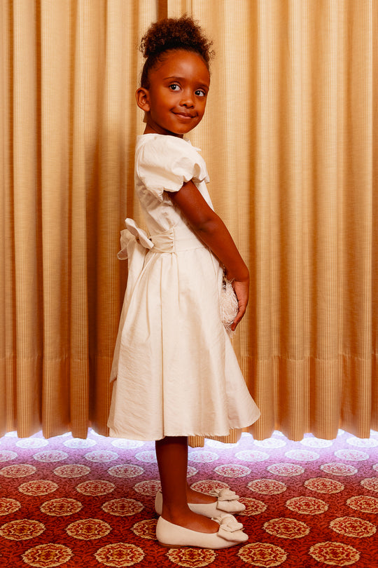 Little girl wearing a white dress standing sideways against a yellow curtain and on a red carpet
