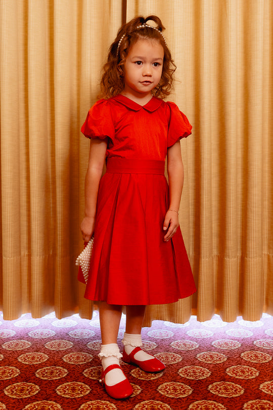 Young girl in a red dress standing in front of a patterned curtain.