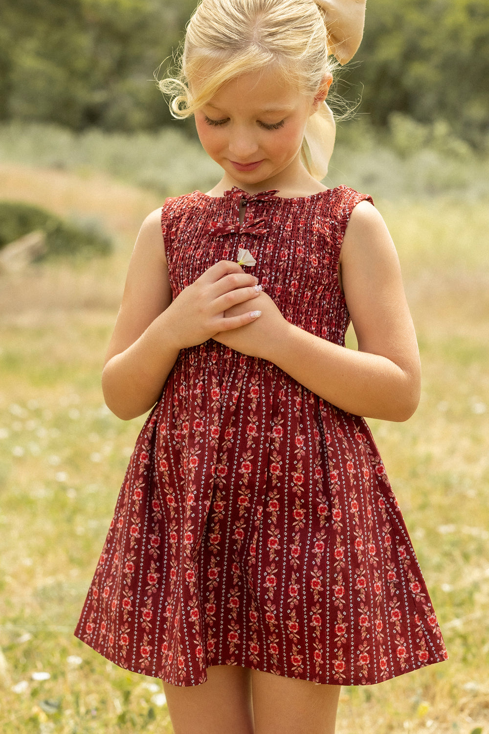 A young blonde girl in the Ivy City Co Mini Ana Dress in Sequoia Felicity Floral stands outside on grass, looking down with her hands together, surrounded by trees and greenery in the blurred background.