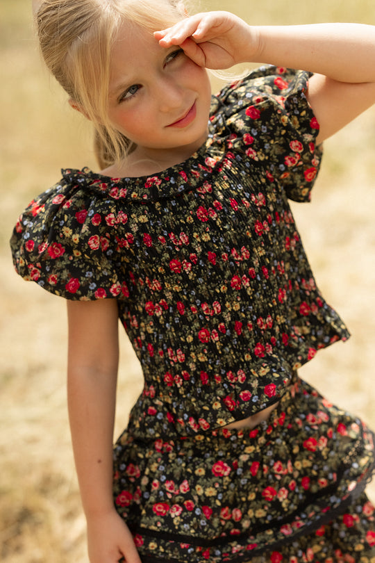 A young girl smiles outdoors in the Ivy City Co Mini Annalise Top in Obsidian Vintage Poppies, shielding her eyes from the sun and enjoying a charming Mom and Me matching moment against a sunny, blurred background.