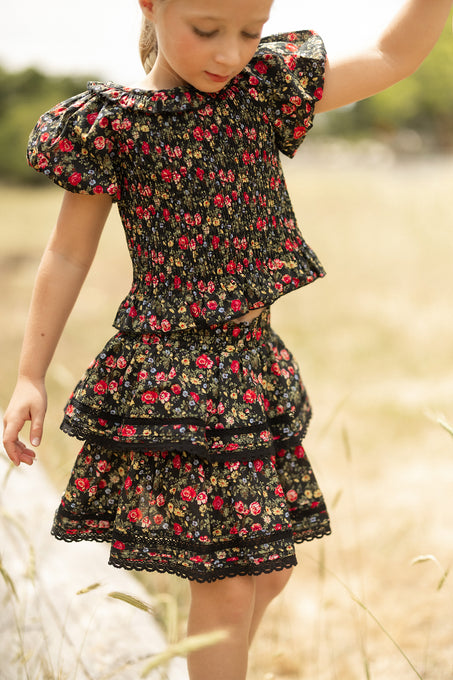 A young girl wears the Ivy City Co Mini Annalise Top in Obsidian Vintage Poppies, standing in a sunlit grassy field with one arm raised, gazing down.