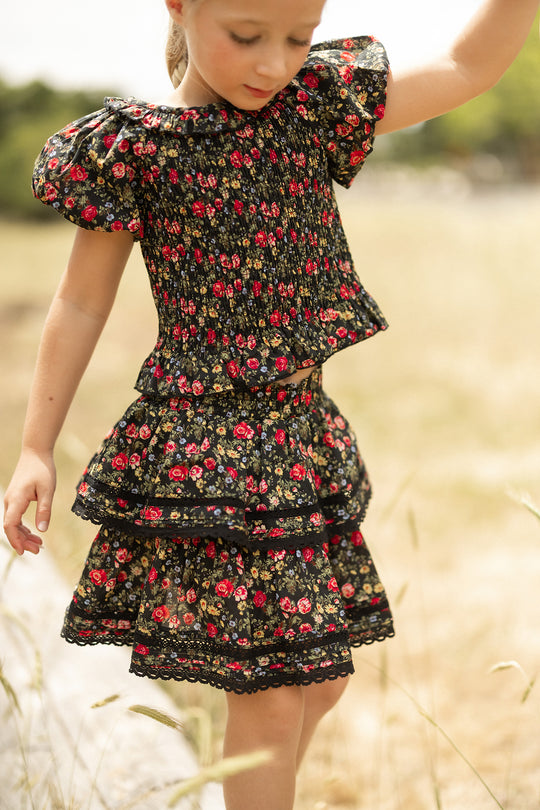 A young girl wears the Ivy City Co Mini Annalise Top in Obsidian Vintage Poppies, standing in a sunlit grassy field with one arm raised, gazing down.