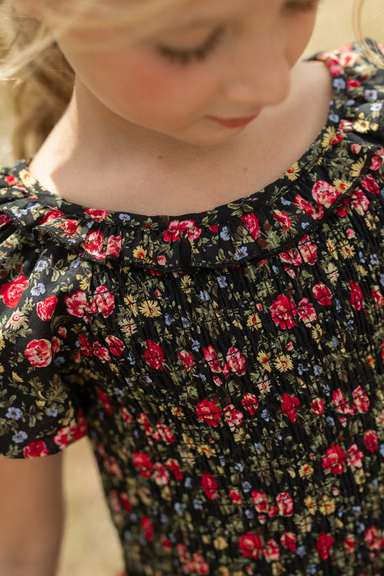 A young child wears the Ivy City Co Mini Annalise Top in Obsidian Vintage Poppies, featuring red, yellow, and blue floral print with ruffled sleeves and neckline. The child's face is partially visible as they look down softly.