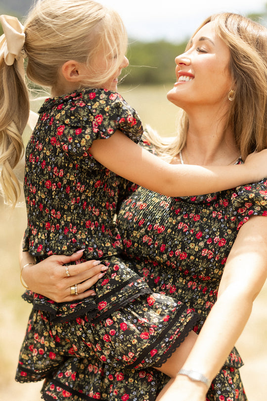 A smiling woman and her daughter enjoy the outdoors, both wearing Ivy City Co’s Mini Annalise Top in Obsidian Vintage Poppies—matching black floral tops that highlight their joyful bond.