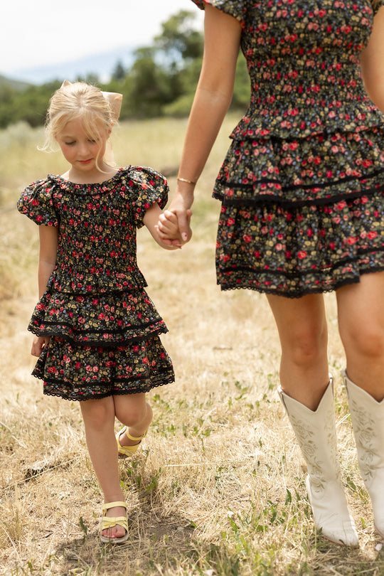 A young girl and an adult hold hands outdoors in matching Ivy City Co Mini Annalise Tops in Obsidian Vintage Poppies, styled with white cowboy boots—Mom and Me matching style amid grass and trees.