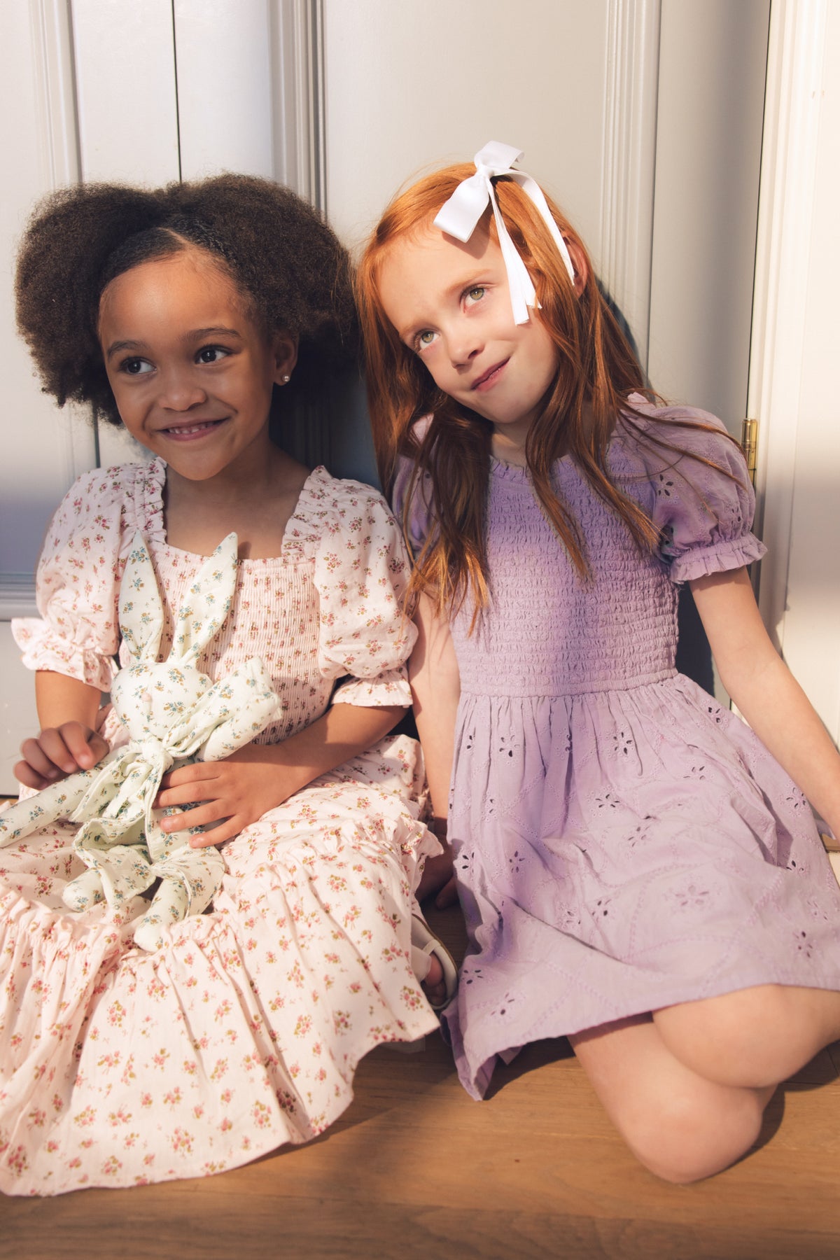 Two young girls sit by a sunlit wall, laughter bright. One wears a floral dress, hugging her toy; the other, with a white bow and long red hair, shines in Ivy City Co’s Mini Emmi Dress in Lilac—perfect for stylish playtime.