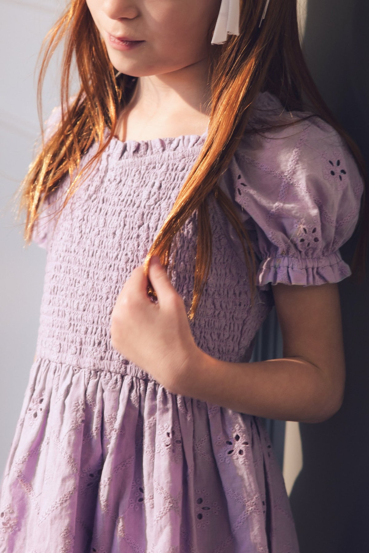A young girl with long reddish hair stands in sunlight, gently touching her hair while wearing the Ivy City Co Mini Emmi Dress in Lilac.