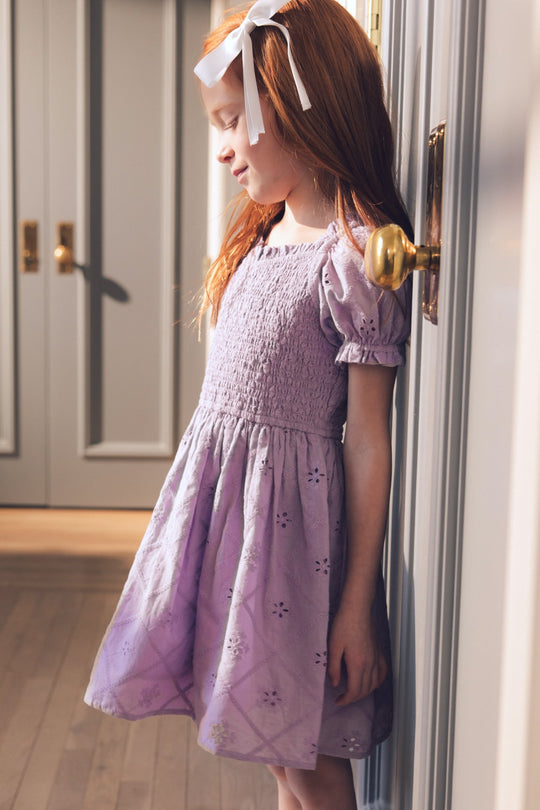 A young girl with long red hair and a white bow stands indoors by a door, softly illuminated by sunlight. She wears the Mini Emmi Dress in Lilac from Ivy City Co—a light spring dress with puffed sleeves—looking down with a gentle expression.