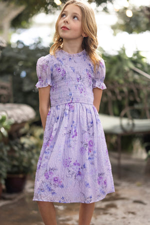 A young girl with blond hair stands outdoors in the Mini Flynn Dress in Lavender Aquarelle Fleur by Ivy City Co, looking up thoughtfully amidst greenery and garden furniture.
