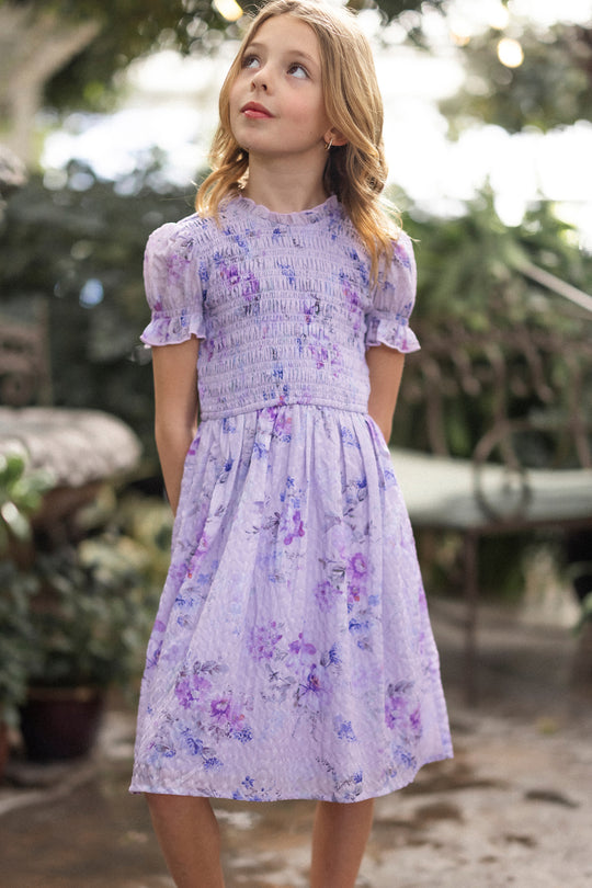 A young girl with blond hair stands outdoors in the Mini Flynn Dress in Lavender Aquarelle Fleur by Ivy City Co, looking up thoughtfully amidst greenery and garden furniture.