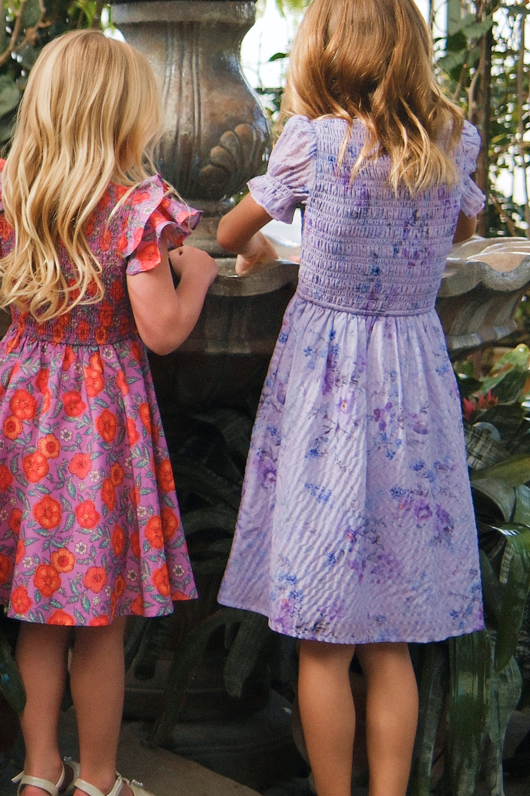 Two young girls, backs to the camera, stand by a stone fountain surrounded by lush greenery. One wears Ivy City Co’s Mini Flynn Dress in Lavender Aquarelle Fleur, while the other wears a watercolor floral dress.