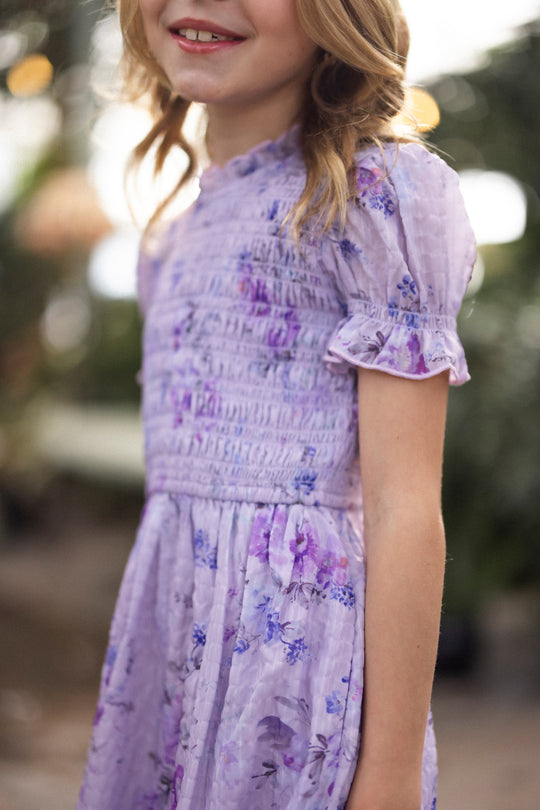 A young girl wearing the Ivy City Co Mini Flynn Dress in Lavender Aquarelle Fleur, with ruffled sleeves and a floral pattern, stands outdoors smiling as the softly blurred greenery and gentle light form a perfect backdrop.