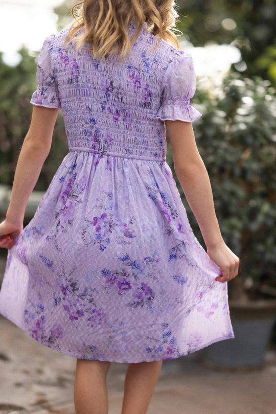 A girl with wavy blonde hair, wearing the Ivy City Co Mini Flynn Dress in Lavender Aquarelle Fleur, stands outdoors facing away and gently holding her dress, with blurred lush green plants in the background.