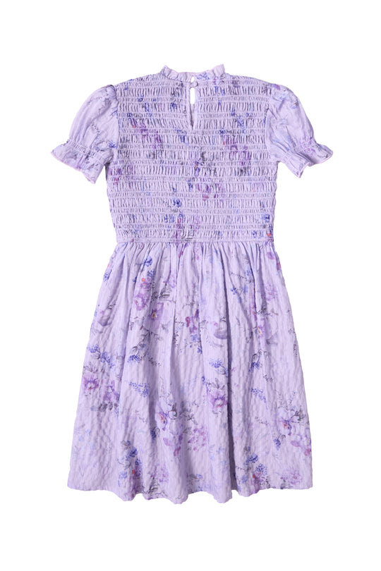 The Ivy City Co Mini Flynn Dress in Lavender Aquarelle Fleur features a smocked bodice, short sleeves with ruffled cuffs, a gently flared skirt, and textured fabric with a watercolor floral print.