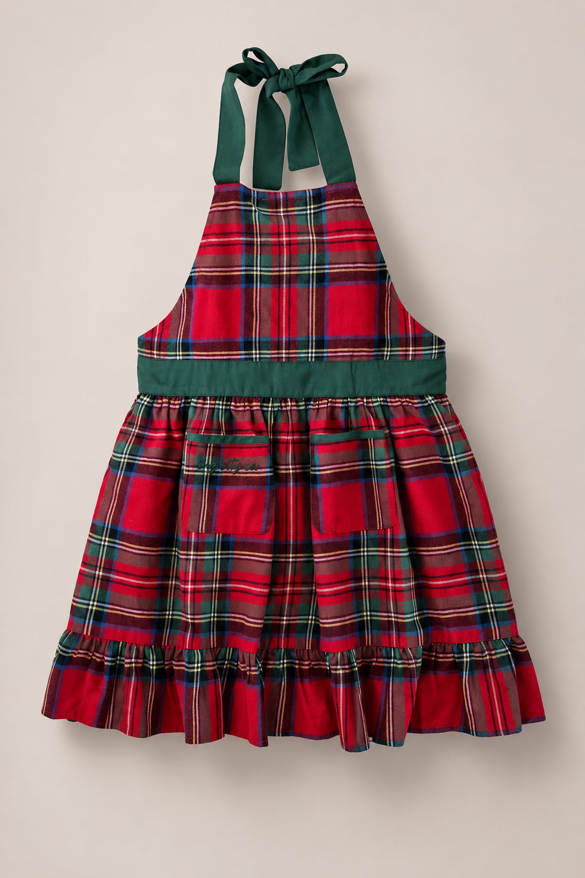 The Ivy Apron in Holiday Plaid by Ivy City Co features a festive red and green plaid pattern, two front pockets, a green waistband, ruffled hem, and green neck ties. Displayed against a plain light background, this apron is the perfect choice for preparing Christmas dinner.