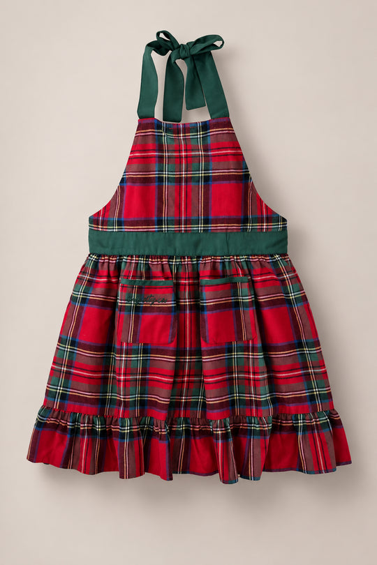 The Ivy Apron in Holiday Plaid by Ivy City Co features a festive red and green plaid pattern, two front pockets, a green waistband, ruffled hem, and green neck ties. Displayed against a plain light background, this apron is the perfect choice for preparing Christmas dinner.