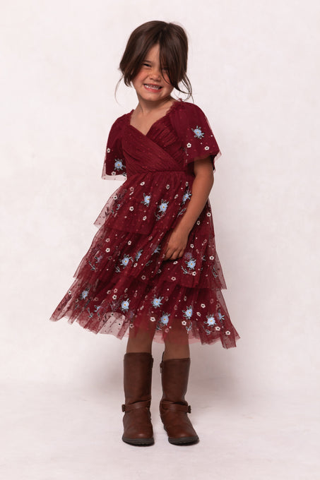 A young girl smiles in the Ivy City Co Mini Mabel Dress in Burgundy, featuring blue florals and a tulle skirt, paired with brown knee-high boots against a plain light background.