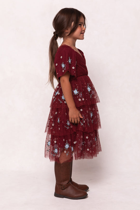 Young girl with long brown hair in a ponytail, wearing the Ivy City Co Mini Mabel Dress in Burgundy with blue floral embroidery and a tiered tulle skirt, plus brown knee-high boots, standing sideways against a light background.
