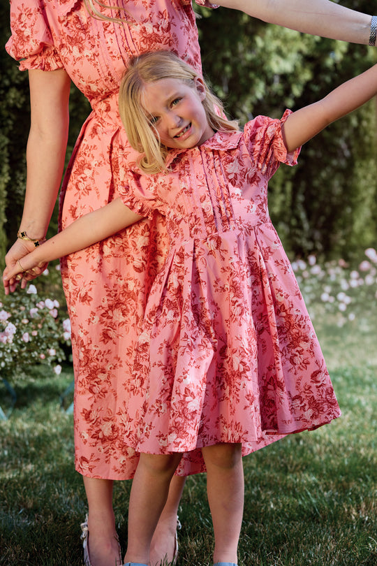 A young girl in the Ivy City Co Mini Sophia Dress in Desert Heritage Rose smiles with arms outstretched as she and an adult, likely her mother, pose outdoors in matching pink floral cotton dresses surrounded by flowers and greenery.