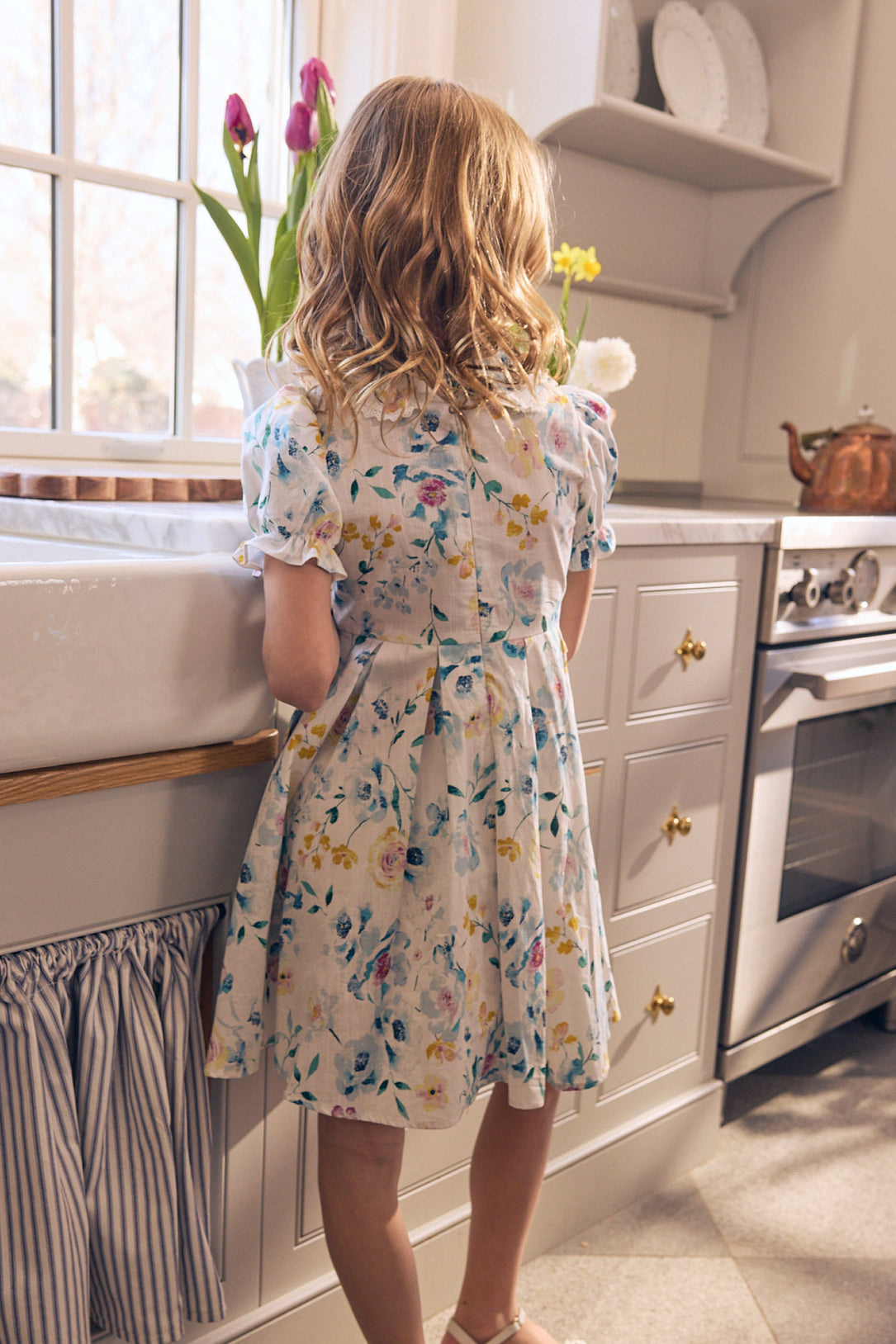 A young girl with wavy blonde hair wearing the Ivy City Co Mini Sophia Dress in Blue Bouquet stands at the kitchen sink, gazing out the window as sunlight streams in and fresh flowers decorate the countertop.