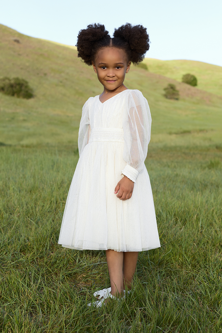 A young girl with curly hair in two puffs stands on a grassy field, wearing the Ivy City Co Mini Stella Dress in White and white shoes, glowing against the green hills and clear sky behind her.