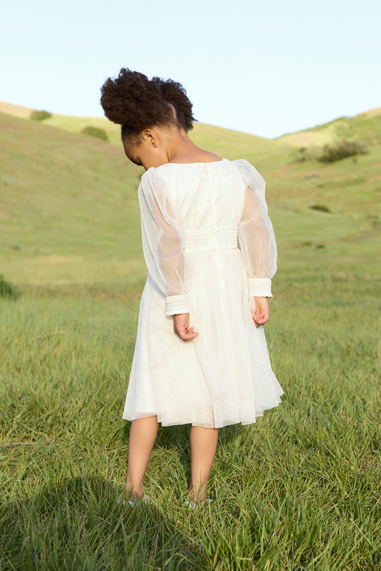 A young girl stands barefoot on grass, looking down, wearing the Ivy City Co Mini Stella Dress in White, with green hills and a clear sky behind her.
