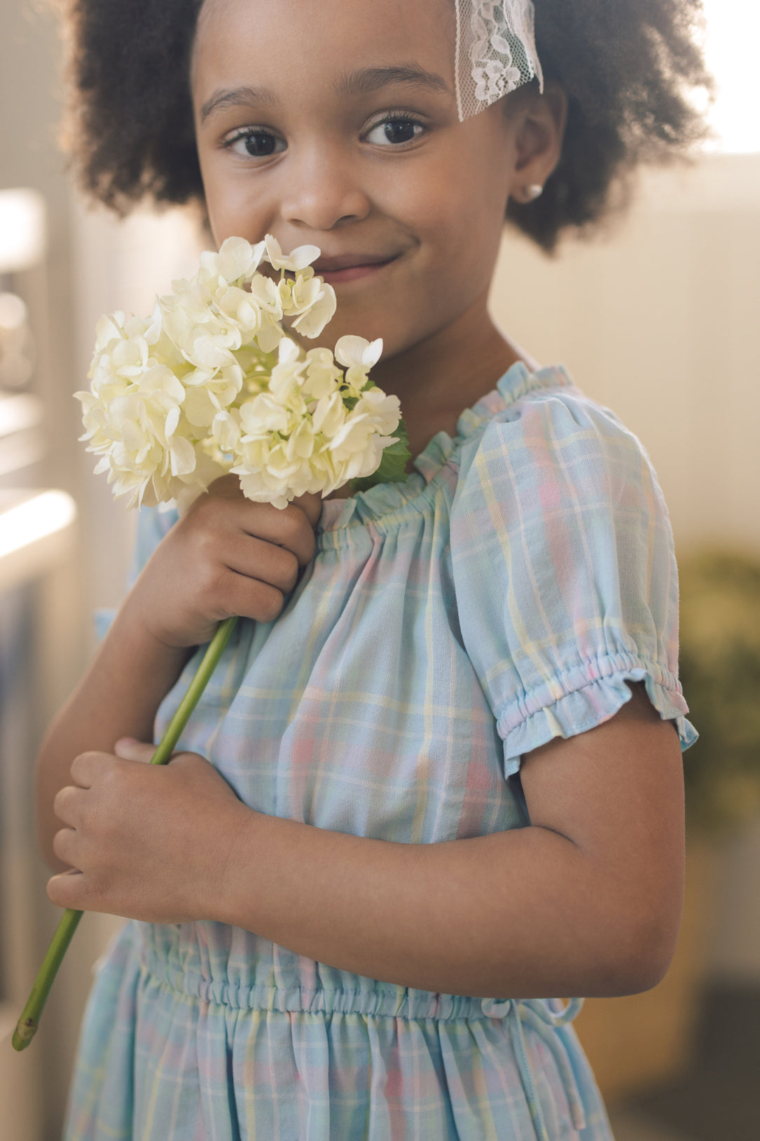 A young girl wears the Ivy City Co Mini Thalia Dress in Pastel Plaid, paired with a lace headband, holding a white hydrangea and smiling softly—perfect for matching mommy and me moments.