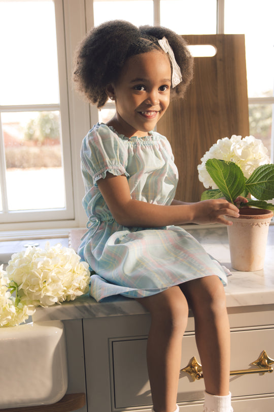 A young girl in the Ivy City Co Mini Thalia Dress in Pastel Plaid sits on a kitchen counter by a window, smiling as she touches a potted plant with white hydrangeas. Sunlight streams through the window behind her.