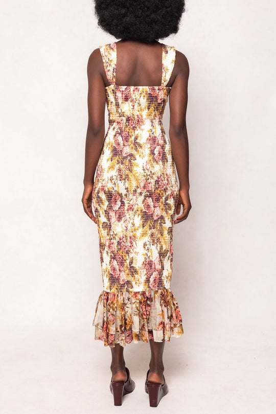A woman with naturally curly hair is shown from the back against a plain background, wearing the Mira Dress in Garden Rose by Ivy City Co—a sleeveless, floral print midi dress with a ruffled hem—paired with brown sandals.