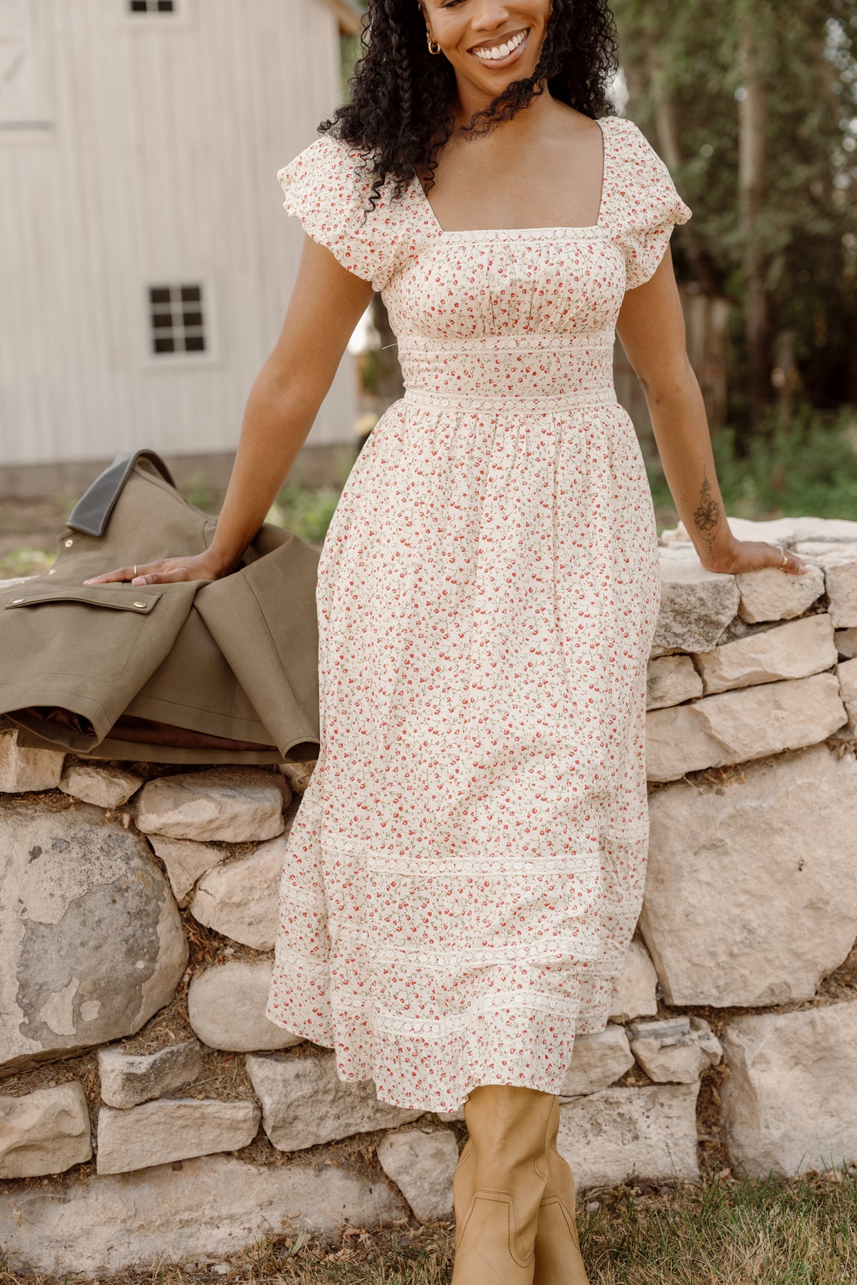 A woman wears the Ivy City Co Mirabelle Dress in Cream Ditsy Floral with puff sleeves and tan boots, smiling as she leans against a stone wall outdoors. A green jacket is draped beside her, with a white barn and trees in the background.