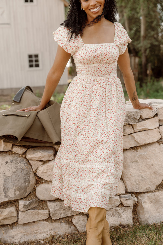 A woman wears the Ivy City Co Mirabelle Dress in Cream Ditsy Floral with puff sleeves and tan boots, smiling as she leans against a stone wall outdoors. A green jacket is draped beside her, with a white barn and trees in the background.