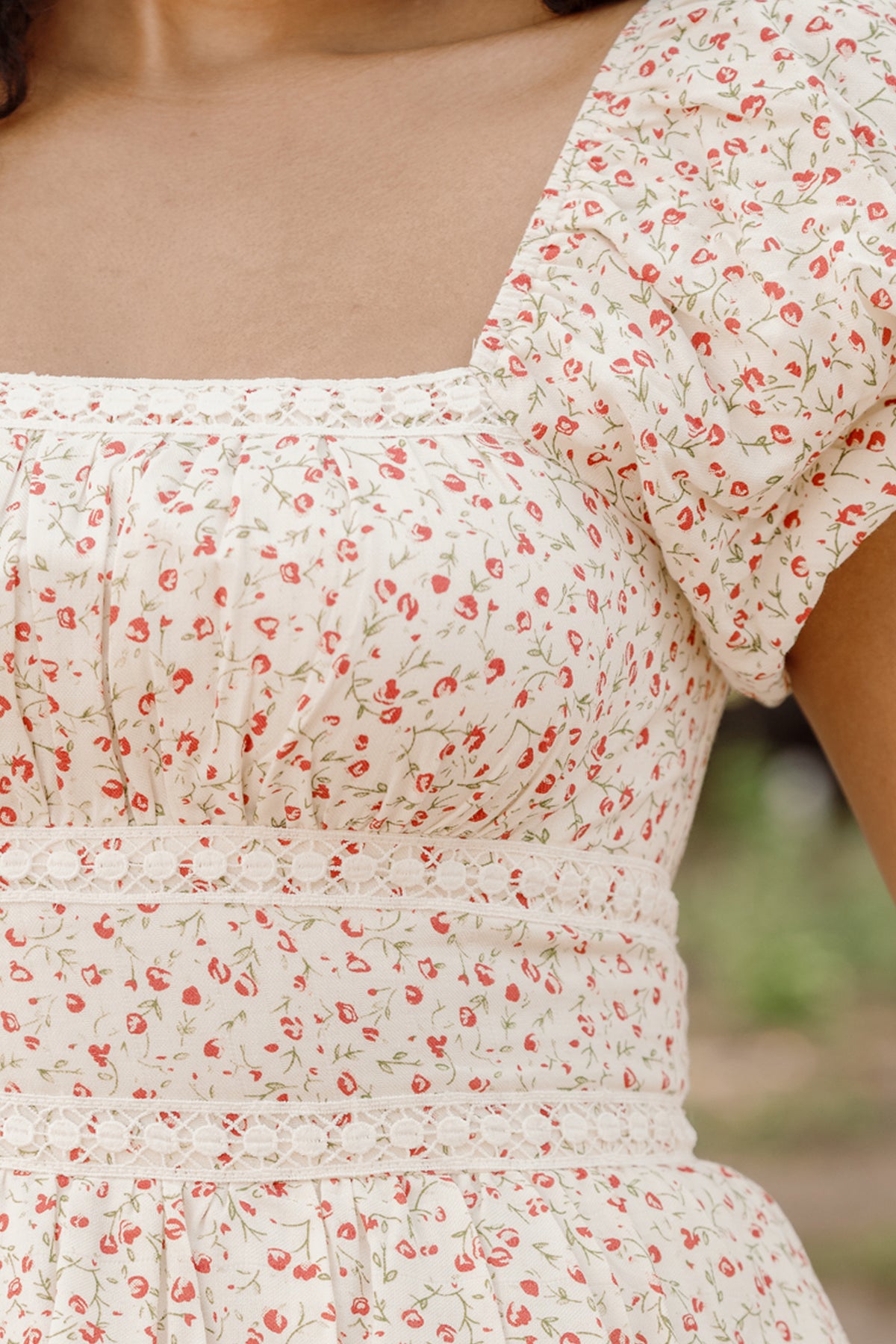 A close-up of a person wearing the Mirabelle Dress in Cream Ditsy Floral by Ivy City Co, featuring short puff sleeves and delicate lace trim at the waist, standing outdoors.
