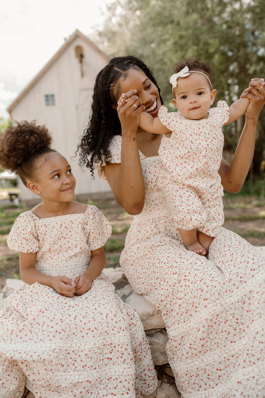 A woman and two young girls sit outside, all dressed in the Ivy City Co Mirabelle Dress in Cream Ditsy Floral, featuring matching white cotton fabric adorned with red floral prints and charming puff sleeves. The woman lifts the baby as the older girl smiles at them, with a barn and trees visible in the background.
