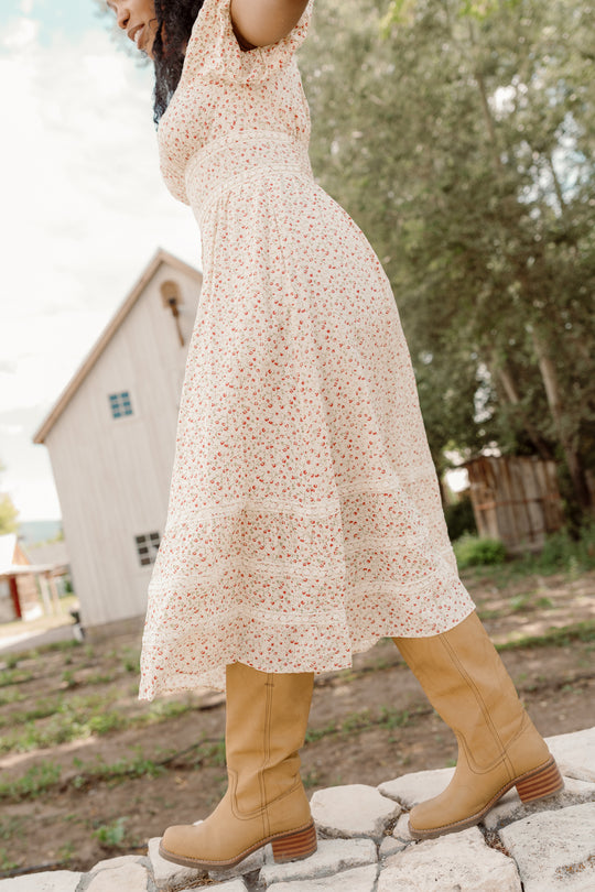 Wearing the Ivy City Co Mirabelle Dress in Cream Ditsy Floral, featuring a smocked bodice and puff sleeves, a person strolls along a stone path with a rustic wooden house and trees behind them. Tan boots complete the charming ensemble.