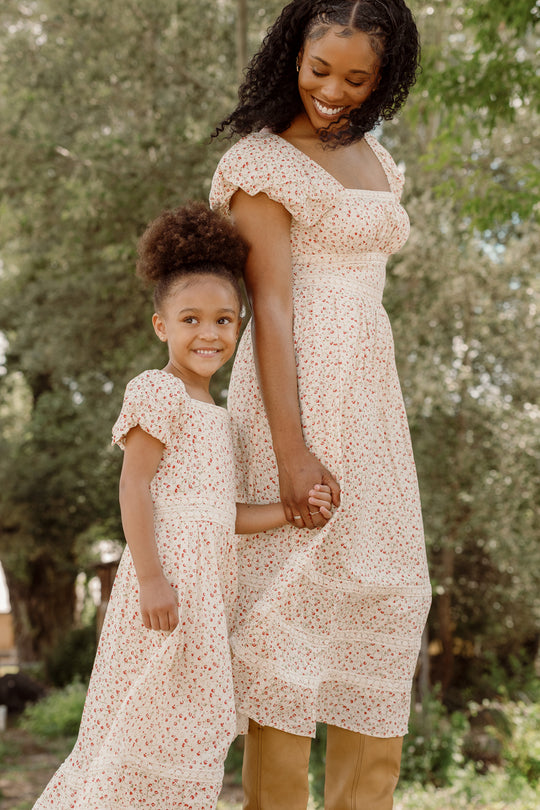 A woman and a young girl, both wearing matching Ivy City Co Mirabelle Dresses in Cream Ditsy Floral with smocked bodices and puff sleeves, hold hands and smile while standing outdoors surrounded by trees.