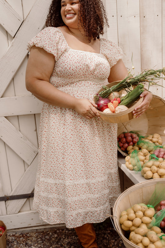 Wearing the Ivy City Co Mirabelle Dress in Cream Ditsy Floral, a woman with puff sleeves stands by baskets of potatoes, smiling and holding a wooden bowl filled with fresh vegetables like zucchini, tomatoes, and onions.