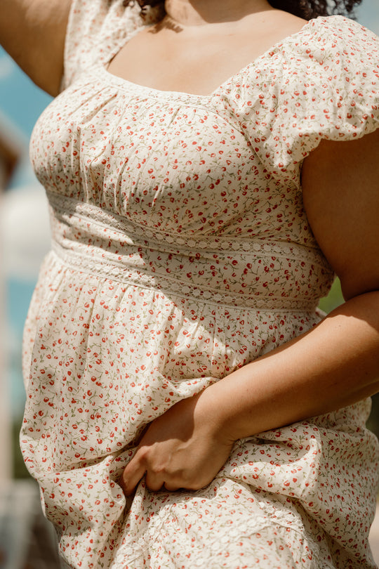 A person wearing the Ivy City Co Mirabelle Dress in Cream Ditsy Floral, featuring a smocked bodice and delicate floral pattern, stands outside holding part of the dress. The photo centers on their torso and arm against a soft blue sky background.