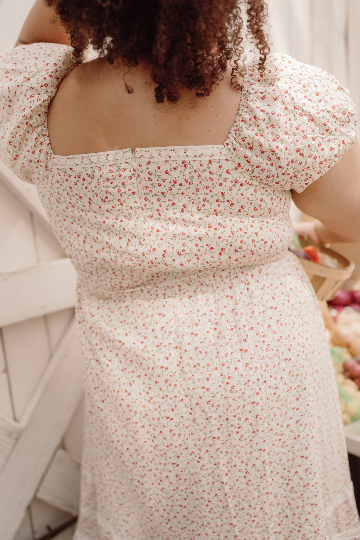 A person with curly hair, wearing the Mirabelle Dress in Cream Ditsy Floral by Ivy City Co—a white cotton dress adorned with a delicate red floral pattern, smocked bodice, and puff sleeves—stands with their back to the camera, holding a basket of fruit.