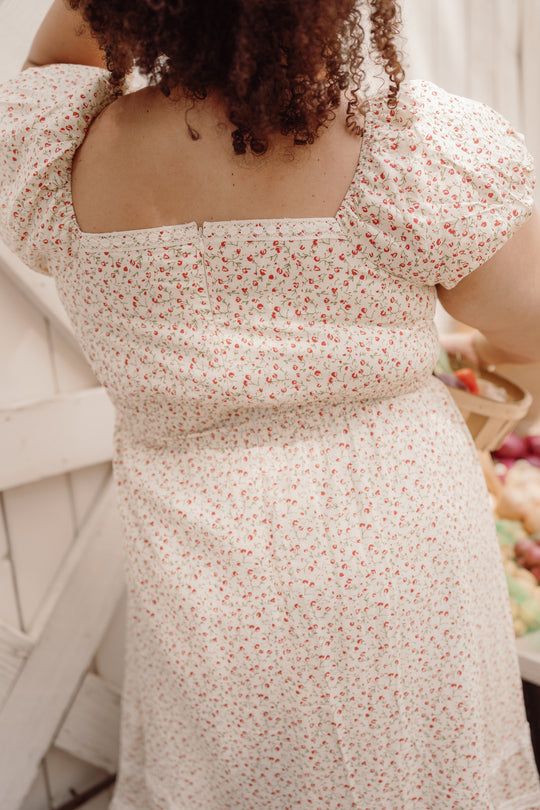 A person with curly hair, wearing the Mirabelle Dress in Cream Ditsy Floral by Ivy City Co—a white cotton dress adorned with a delicate red floral pattern, smocked bodice, and puff sleeves—stands with their back to the camera, holding a basket of fruit.