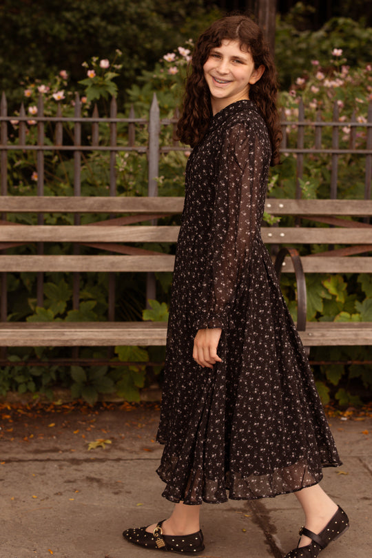 A young woman with long, curly brown hair smiles as she walks past a wooden bench and metal fence, surrounded by green foliage and pink flowers, wearing the Mini Sophia Long Sleeve Dress in Noir Ditsy Chiffon from Ivy City Co paired with black shoes.