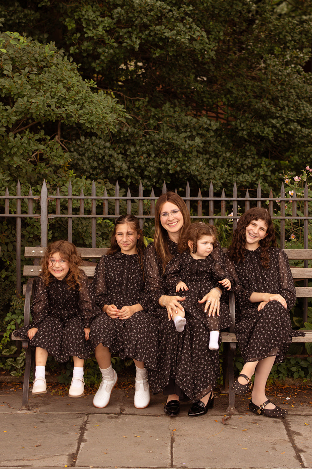 A woman and four young girls sit on a bench, all smiling in their matching Ivy City Co Mini Sophia Long Sleeve Dresses in Noir Ditsy Chiffon. Their coordinated look adds a charming touch to the scene, set against a metal fence and lush greenery.