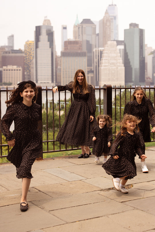 Five girls in coordinating dark dresses, including the stylish Mini Sophia Long Sleeve Dress in Noir Ditsy Chiffon by Ivy City Co, pose and play together on a paved area bordered by a metal fence, with a city skyline and tall buildings visible in the background. The atmosphere is joyful and lively.